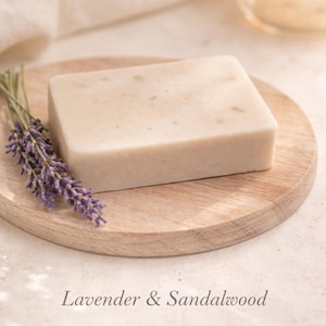 Puede incluir: Una pastilla de jabón rectangular de color claro descansa sobre un plato de madera pequeño, junto a un manojo de lavanda morada. Las palabras "Lavender & Sandalwood" están impresas debajo. Una vela encendida está en el fondo.