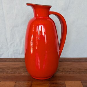 May include: A bright orange ceramic pitcher with a glossy finish, featuring a rounded body, a curved handle on the right side, and a small pouring spout at the top. The pitcher has a smooth, solid-colored surface with a reflective sheen.