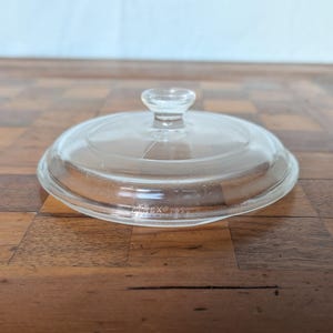 May include: A clear, circular glass lid featuring a small, centered knob handle. The lid has a stepped rim design and includes the embossed text PYREX along the edge, indicating it is a replacement cover for a glass cookware vessel.