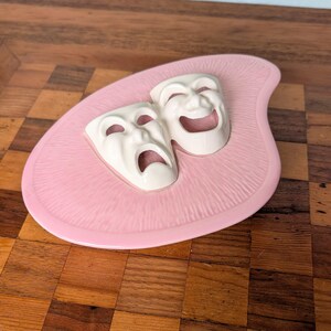 May include: A pink ceramic dish with a textured surface featuring two white theatrical masks in relief. One mask depicts a sad expression with a downturned mouth, while the other mask displays a joyful expression with an upturned, smiling mouth.