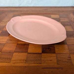 Puede incluir: Una fuente ovalada de cerámica en color rosa claro con un acabado liso y mate. La fuente presenta un borde ligeramente elevado y detalles circulares sutilmente grabados en los lados izquierdo y derecho, creando un diseño sencillo.