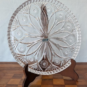 May include: A clear, circular glass serving plate featuring an intricate cut-glass pattern of radiating lines and starburst motifs. The plate has a scalloped, serrated edge and a small rectangular sticker in the center with the text Violetta.