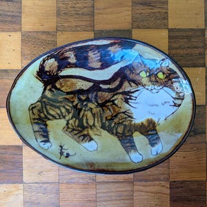 May include: Oval ceramic dish with a hand-painted cat design. The cat is depicted in brown and tan tones with white accents and green eyes. The dish has a light yellow-green background and a dark brown rim. A small, stylized insect is near the cat's paws.