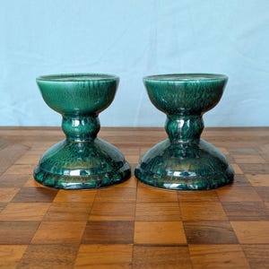 May include: Two matching green ceramic candle holders. Each has a wide, shallow bowl at the top, a rounded stem, and a flared base. The glossy finish has a textured, mottled appearance. The candle holders are displayed on a wooden surface.