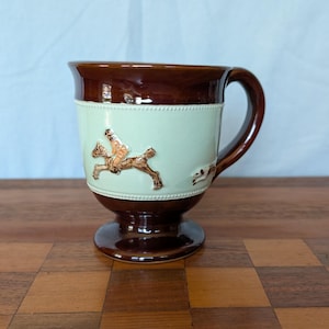 May include: A brown and cream-colored ceramic mug with a handle and pedestal base. The mug features a raised relief design of a rider on a horse and a dog. The top and base are a darker brown, with a cream-colored band around the middle.