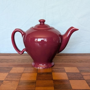 May include: A burgundy ceramic teapot with a lid and a curved handle. The teapot has a spout and sits on a wooden surface. The teapot is a solid color and has a glossy finish.