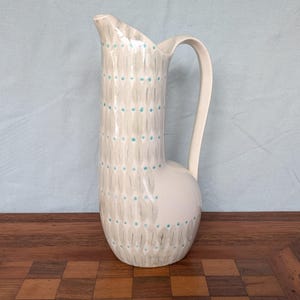 May include: A tall, slender ceramic pitcher with a cream-colored base, featuring a repeating pattern of vertical gray brushstrokes and small, evenly spaced turquoise dots. The vessel has a curved handle and a distinctively shaped pouring spout.