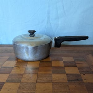 May include: A small, vintage aluminum saucepan with a lid and a black handle. The pot is silver-colored with a dark knob on the lid. The handle is black and attached to the side. The saucepan is sitting on a wooden surface.