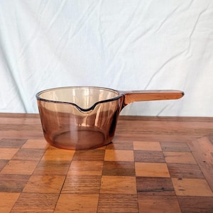 May include: A vintage amber-colored saucepan with a single handle. The glass pot has a pouring spout and is set on a wooden surface. The handle is an extension of the pot's body. The background is a white sheet.