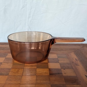 May include: A translucent amber-colored glass saucepan with a round body and a single integrated handle extending from the side. The cookware features a smooth, polished finish and a wide, open top, showcasing a consistent brown tint throughout.