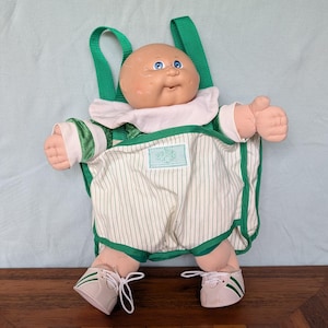 May include: A Cabbage Patch Kids doll with a bald head, blue eyes, and a round face. The doll wears a white collared shirt with green sleeves, white pinstriped overalls with a green trim, and white sneakers with green stripes and white laces.