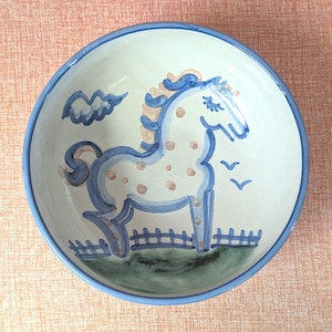 May include: A white ceramic bowl with a blue rim and a hand-painted horse design. The horse is blue with pink spots and a pink mane, set against a blue fence and green grass. A blue cloud and birds complete the scene.