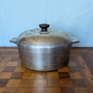 May include: A vintage silver-colored cast iron Dutch oven with a lid and a dark brown knob. The pot has two side handles and sits on a wooden surface with a checkerboard pattern. The background is a solid light blue.