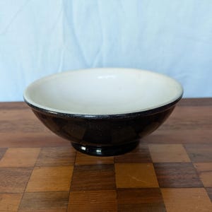 May include: A small, round bowl with a black exterior and a white interior. The bowl has a slightly flared rim and sits on a small, circular base. The bowl is on a wooden surface.