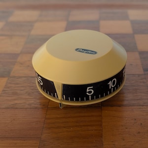 May include: A round, beige kitchen timer with a sloped top and a black side band featuring white numerical markings from 5 to 55. The word Dayton is printed in a blue oval on the top surface. The device has a small vertical indicator pointer.