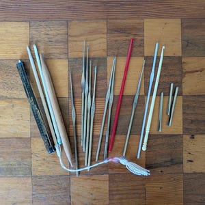 May include: A collection of crochet hooks in various sizes and materials, including wood, metal, and plastic. The hooks are arranged on a checkered wooden surface, with a white tassel and a black measuring tool.