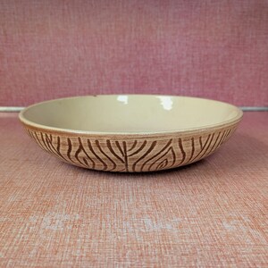 May include: A ceramic bowl with a cream-colored interior and a brown, textured exterior. The bowl's exterior features a repeating pattern of curved lines, creating a leaf-like design. The bowl is set against a pink background.