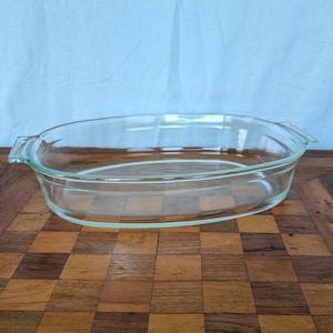 May include: Clear, oval-shaped glass baking dish with handles. The dish is empty and sits on a wooden surface with a checkered pattern. The dish is designed for baking and serving.
