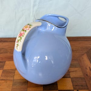 May include: A light blue ceramic pitcher with a rounded body and a glossy finish. The handle is cream-colored and features a delicate floral pattern with pink roses and green leaves. The rim of the pitcher has a thin, dark metallic trim.