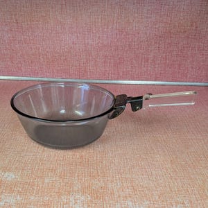 May include: A clear glass saucepan with a black rim and a clear plastic handle. The handle is attached to the pan with a metal bracket. The pan is sitting on a textured, peach-colored surface.