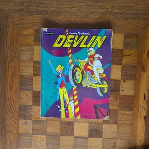 May include: A vintage Hanna-Barbera's Devlin coloring book featuring a purple cover with a yellow title. The illustration depicts a character on a motorcycle performing a stunt, alongside two other figures, set against a geometric background.