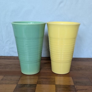 May include: Two tapered drinking tumblers with a ribbed texture along the exterior. One tumbler is a soft pastel green, and the other is a bright yellow. Both vessels feature a smooth, glossy finish and a wide, open rim, standing side by side.