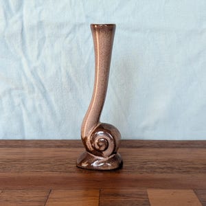 May include: A tall, slender ceramic bud vase with a glossy brown speckled glaze. The base features a distinct, rounded spiral or snail-like shape that curves upward into a long, narrow neck with a small circular opening at the very top.