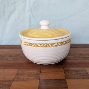 May include: A round ceramic sugar bowl with a speckled off-white base and a yellow lid. The bowl features a horizontal band with a repeating geometric triangle pattern in yellow. The body has subtle horizontal ridges and a matching yellow knob.