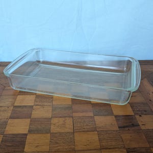 May include: A rectangular, clear glass baking dish with rounded corners and handles. The dish is empty and sits on a checkered wooden surface, with a light blue backdrop.