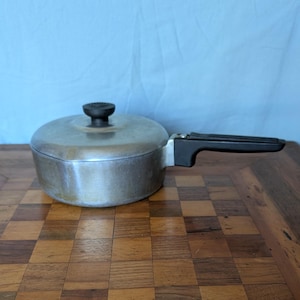 May include: A vintage silver-toned saucepan with a black handle and lid. The lid has a black knob. The pan is sitting on a wooden checkerboard surface. The background is a light blue fabric.
