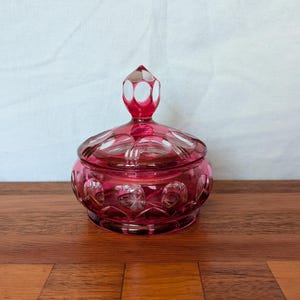 May include: A ruby-red, lidded glass jar with a faceted design. The jar has a round base and a lid with a decorative finial. The glass is cut with a pattern of circles and ovals. The jar is sitting on a wooden surface.
