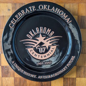 May include: A round black commemorative plate featuring copper-colored embossed text and graphics. The text reads CELEBRATE OKLAHOMA! OKLAHOMA CENTENNIAL 1907-2007 A UNIQUE HISTORY. AN EXTRAORDINARY FUTURE. The center displays a star and the number 07.