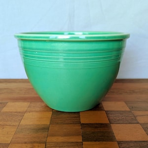 May include: A vintage, green ceramic bowl with a rounded shape and a slightly flared rim. The bowl features horizontal, etched lines around the middle. The bowl is a vibrant, solid green color and sits on a wooden surface.