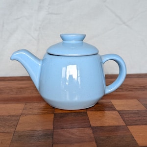 May include: A light blue ceramic teapot with a speckled finish, featuring a rounded body, a curved spout, a matching lid with a small knob handle, and a side handle. The teapot has a smooth, glossy surface that reflects light on its side.