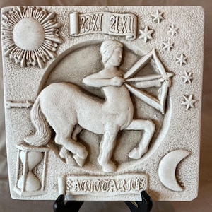 Puede incluir: Placa del signo zodiacal Sagitario de color crema. La placa presenta un relieve de un arquero centauro, estrellas, una luna creciente, un sol y un reloj de arena. El texto "SAGITTARIUS" está debajo del centauro. Las fechas "23XI 21XII" están arriba.