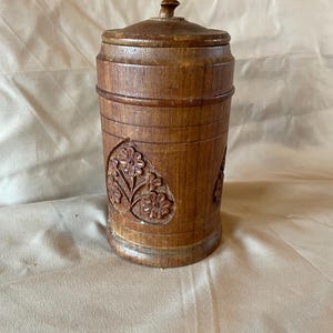 Puede incluir: Un recipiente cilíndrico de madera con tapa, con diseños florales tallados. El recipiente es de color marrón cálido, con un aspecto ligeramente envejecido. La tapa tiene un pequeño pomo en la parte superior. El recipiente se utiliza probablemente para el almacenamiento.