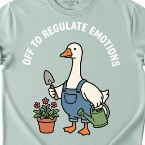 May include: A light green t-shirt featuring a cartoon goose wearing blue overalls, holding a trowel and a watering can. A potted plant with red flowers is to the left. The text "OFF TO REGULATE EMOTIONS" is arched above the goose.