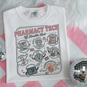 May include: White t-shirt with a colorful graphic design. The design features the text "PHARMACY TECH Health Hub" in pink, along with illustrations of pharmacy-related processes and equipment. The shirt is a crew neck style.