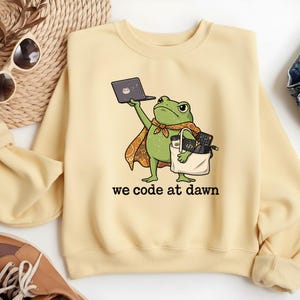 May include: A light yellow crewneck sweatshirt featuring a graphic of a green frog wearing a patterned cape. The frog holds a laptop in one hand and a tote bag filled with tech accessories in the other. The text reads, we code at dawn.