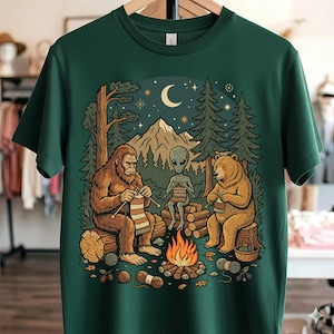 May include: Dark green t-shirt featuring a whimsical illustration of a Bigfoot, an alien, and a bear knitting around a campfire in a forest setting. The design includes a mountain range, trees, a crescent moon, and stars.