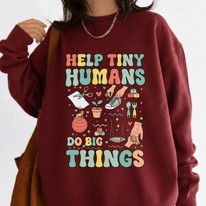 Puede incluir: Sudadera burdeos con el texto "HELP TINY HUMANS DO BIG THINGS" en letras coloridas de estilo retro. El gráfico incluye ilustraciones de tijeras, una planta, un zapato y otros elementos lúdicos.