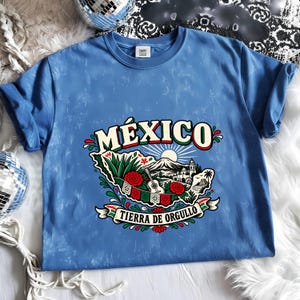 May include: A blue acid-wash t-shirt featuring a graphic of Mexico with the text "MÉXICO" and "TIERRA DE ORGULLO". The design includes a sun, a mountain, a guitar, red flowers, green leaves, and a banner in red, white, and green colours.