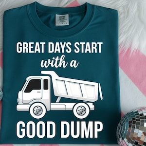 May include: Teal t-shirt with the text "GREAT DAYS START with a GOOD DUMP" in white. A white dump truck graphic is below the text. The shirt is folded and displayed on a pink and white striped surface.