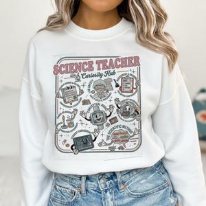 Puede incluir: Sudadera blanca con un gráfico colorido que dice "SCIENCE TEACHER Curiosity Hub" en rosa y verde. El gráfico incluye ilustraciones sobre seguridad en el laboratorio, cuestionarios y el método científico. La sudadera es de cuello redondo.
