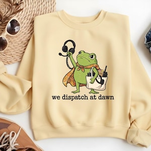 May include: A light yellow crewneck sweatshirt featuring a graphic of a green frog wearing a patterned cape, holding a headset in one hand and a white tote bag containing a radio and pens in the other, with the text we dispatch at dawn below.