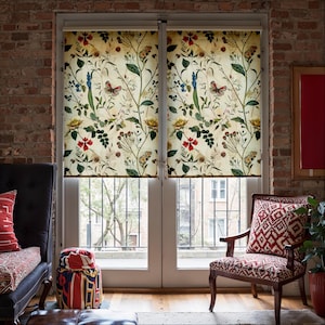 Op de afbeelding: Raamrolgordijnen met een bloemenpatroon met vlinders, rode, gele en blauwe bloemen en groene bladeren. De rolgordijnen staan tegen een bakstenen muur en een raam.