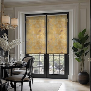 May include: Two window roller blinds featuring a yellow backdrop with a floral and bird design. The blinds are installed in a room with black-framed doors and a view of an outdoor patio. A dining table and chairs are in the foreground.