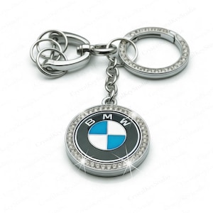 Pode incluir: Um porta-chaves BMW prateado com um emblema redondo com o logótipo da BMW em azul, branco e preto, rodeado por cristais brilhantes. O porta-chaves inclui uma corrente, um porta-chaves e um fecho.