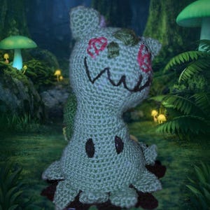 May include: Crocheted plush toy in the shape of a ghost-like creature. The toy is primarily light green with dark brown accents and pink floral details. It has a smiling mouth and two dark brown oval eyes. The plush has a whimsical, handmade appearance.