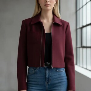 May include: A burgundy cropped jacket with a collared neckline and a silver zipper. The jacket has a single pocket and is paired with blue jeans and a black top. The model is standing in front of a window.